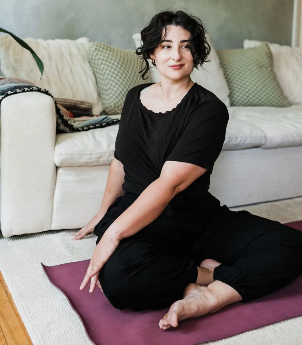A brown haired woman sitting on the floor with crossed legs in a twist. She smiles gently with warmth.