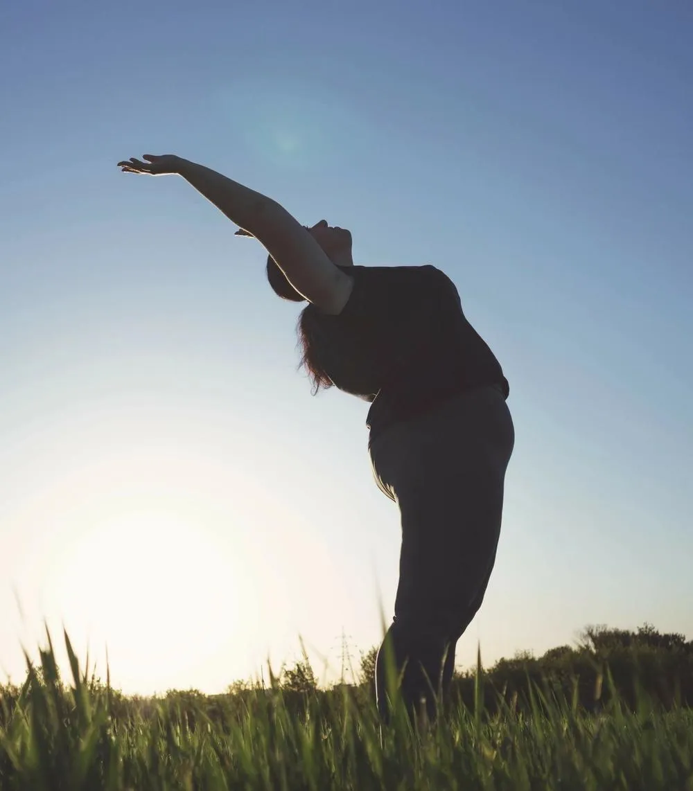 An average-bodied woman stands in a field of grass with her hands lifted to wards the sky.