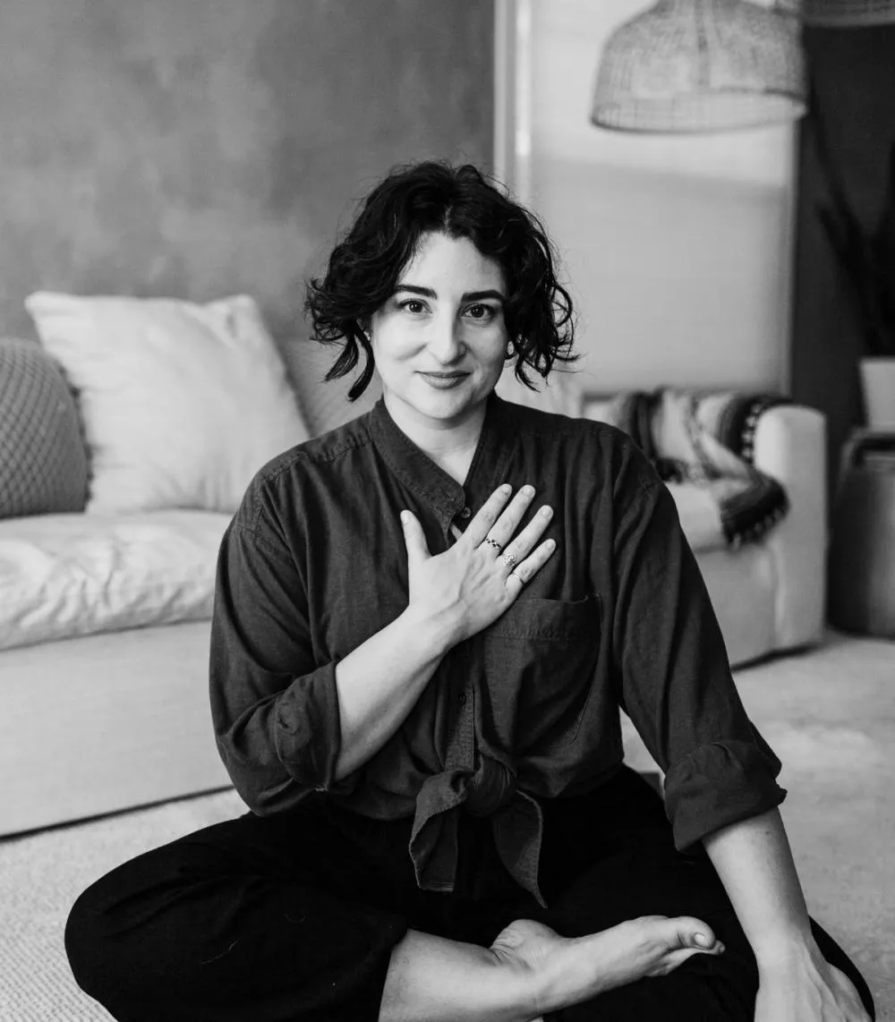 A brown haired woman sitting on the floor with crossed legs. Her hand is over her heart and she smiles gently with warmth.