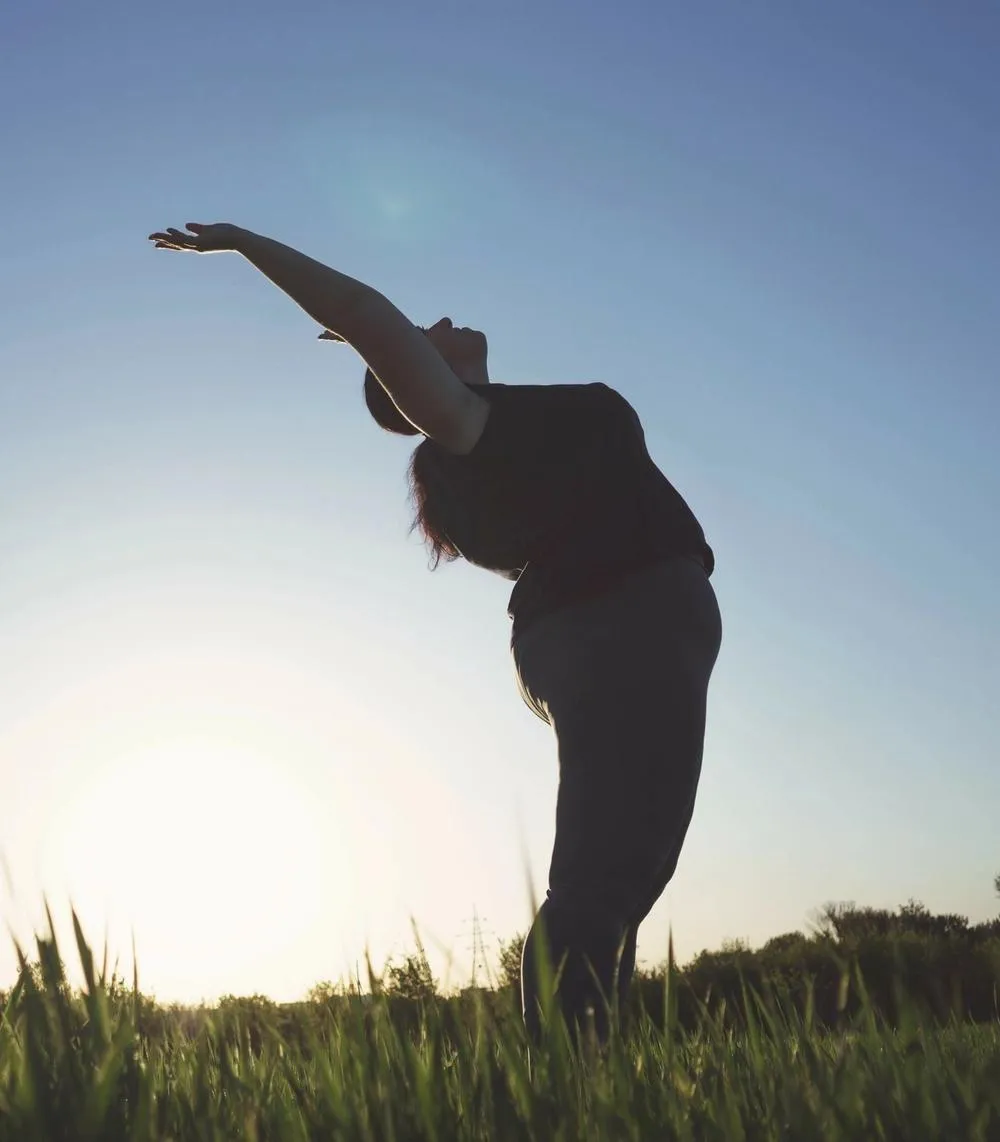 An average-bodied woman stands in a field of grass with her hands lifted to wards the sky.